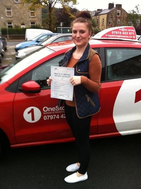 Learner holding a practical driving test pass certificate beside the red One Stop School of Motoring car in Burnley